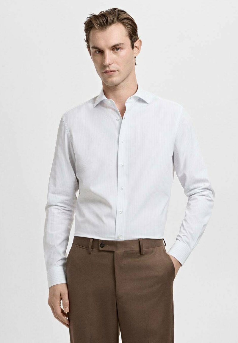 Chemise habillée blanche à manches longues avec des rayures verticales, col classique, devant à boutons, associée à un pantalon marron, texture de tissu lisse.