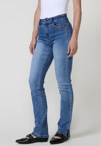 Sinivärvilised teksad kitsase lõikega, kõrge vöökohaga ja kaunistatud eesmistest taskutest. Kangas on sileda tekstuuriga ja kergelt pestud effekiga.