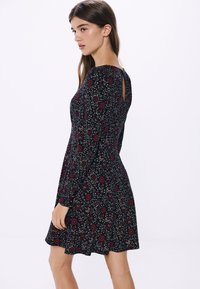 Svart klänning med blommönster och långa ärmar, med en nyckelhålrygg och en utsvängd kjol, tillverkad av mjukt material.