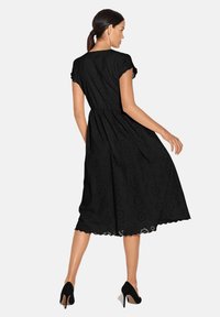 Schwarzes midikleid aus Lochstoff, mit tailliertem Schnitt und gewelltem Saum. Kurze Ärmel und lockere Silhouette. Schwarze High Heels.