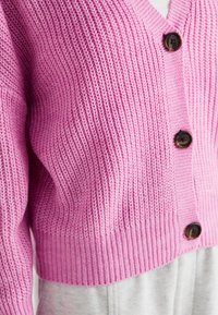 Stickad rosa cardigan med V-ringning, strukturerad mönster och fem bruna knappar. Slutar vid midjan med ribbade manschetter.