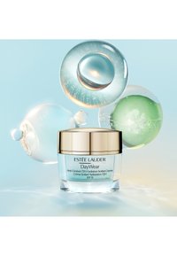 Estee Lauder DayWear-krämburk i ljusblått glas med gulddetaljer, med lock, omgiven av färgstarka, cirkulära geléliknande element på en mjuk bakgrund.