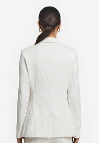 Vit mönstrad kavaj med en texturerad tyg. Har en enkel slits, snedskurna slag och slät foder. Framhäver en rak silhuett.