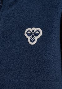 Sjöblå fleece tyg med en strukturerad yta. Inkluderar en vit broderad logotyp med sjöblå accenter på vänster sida.