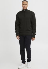 Ein Mann steht mit Blick nach vorne und trägt einen dunkel strukturierten Halbzip-Pullover, schwarze schmale Hosen und weiße Sneakers vor einem schlichten hellen Hintergrund.