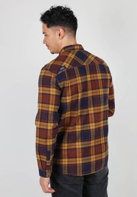 Chemise en flanelle à carreaux marron et jaune avec des accents bleus ; dotée de manches longues, d'une patte de boutonnage et d'un logo sur l'épaule.