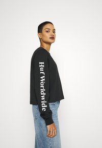 Fekete, hosszú ujjú póló, amelyen fehér színnel függőlegesen "Huf Worldwide" felirat található a bal ujján, világoskék, egyenes szárú farmernadrággal viselve.