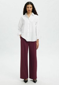 Femme debout les mains dans les poches, portant une chemise blanche boutonnée, un pantalon large bordeaux et des chaussures noires pointues sur fond uni.