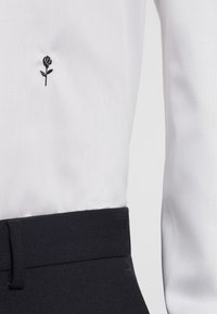 Chemise blanche avec un motif de rose brodé noir sur le côté gauche, associée à un pantalon en tissu texturé foncé.