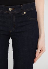 Mörkblå denimjeans med guldfärgad metall, kontrasterande sömmar, en klassisk femficksdesign och en åtsittande silhuett.