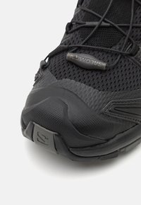 Scarpa sportiva nera con tomaia in mesh, punta in gomma, dettagli sintetici testurizzati e lacci a bungee. Logo ben visibile sul lato.