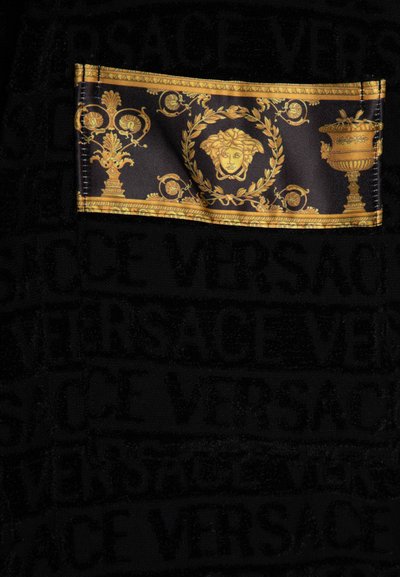 Tissu noir présentant des logos "Versace" en relief et audacieux. Un patch accentué de doré arbore des designs complexes, y compris un visage et des motifs ornés.
