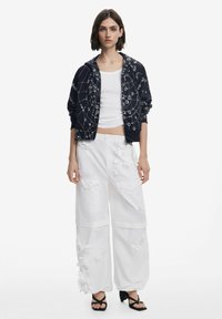 Chaqueta navy con cremallera y patrón de telaraña con detalles plateados; pantalones blancos de pierna ancha con apliques florales texturizados; sandalias negras de tacón.