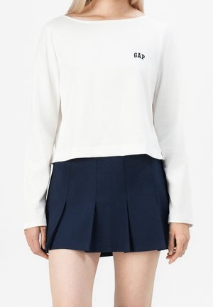 Vrouw draagt een wit shirt met lange mouwen met "GAP"-logo en een marineblauwe geplooide minirok, staand tegen een witte achtergrond.