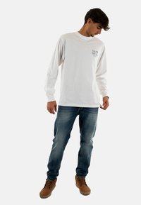 Camisa blanca de manga larga con un diseño impreso en negro; combinada con jeans de mezclilla azul. El modelo lleva botas de cordones marrón claro.