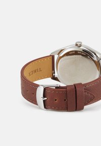 Cinturino in pelle marrone con fibbia in acciaio inox argentato. Il retro dell'orologio presenta dettagli incisi. Texture liscia e design leggermente curvato.
