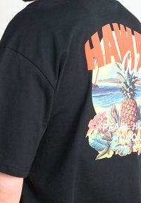 Černé tričko s barevným potiskem ve stylu Hawaii, obsahující ananas, tropické květiny, oceán a výrazný oranžový text "HAWAII".