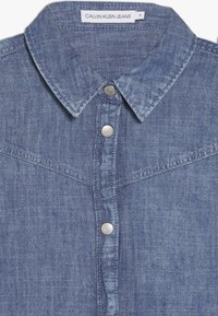 Světle modrá denimová košile s límcem na knoflíčky, stříbrnými patentkami a detaily západního stylu. Textura látky je mírně opotřebovaná.