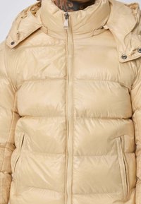 Doudoune beige avec zip frontal et poches latérales, portée par une personne avec un tatouage visible sur le cou.