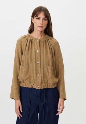 Mujer con pelo liso y castaño que lleva una chaqueta camel abotonada con bolsillos delanteros y pantalones azul oscuro de talle alto sobre un fondo blanco.