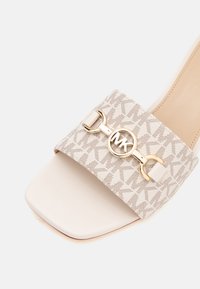 Sandale beige à glissière avec une large bride présentant un motif MK répété et une boucle circulaire dorée avec le logo MK sur le dessus.