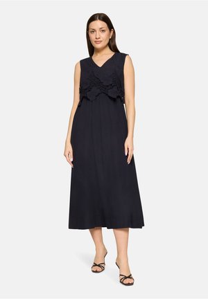 Vrouw draagt een mouwloze zwarte midijurk met bloemige kanten overlay op het lijfje en zwarte sandaaltjes met bandjes, staand voor een effen achtergrond.