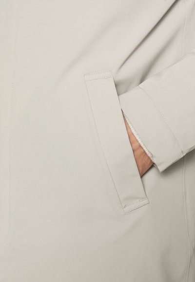 Abrigo beige con una textura suave, que presenta un bolsillo lateral y un puño de muñeca ligeramente acolchado. Detalles de costura visibles a lo largo de la tela.