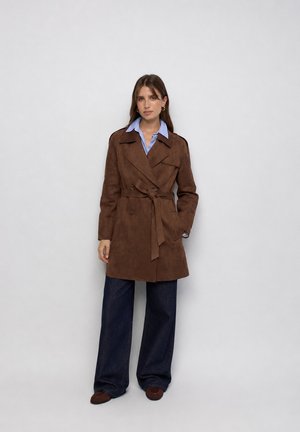 Mujer con un abrigo trench marrón con cinturón sobre una camisa azul con cuello y vaqueros anchos y oscuros, de pie frente a un fondo claro y simple.