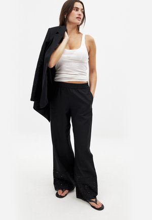 Pantaloni neri a gamba larga con fondo strutturato caratterizzato da fori a occhio, abbinati a un top bianco senza maniche e a un blazer nero appoggiato sul braccio.