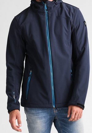 Homme portant une veste softshell bleu marine avec une fermeture éclair bleu sarcelle et des poches zippées, associée à un jean bleu clair.
