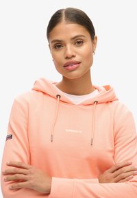 Jasnopomarańczowy hoodie z kapturem i sznurkiem; posiada białe logo "SUPERDRY"; miękka, casualowa tkanina; długie rękawy z detalami w formie łat. 