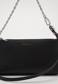 Pochette en cuir noir avec une surface texturée, équipée d'une fermeture zippée, d'une bandoulière en chaîne et d'un logo "HUGO" en relief à l'avant.