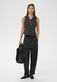 Tmavošedý bezrukávový sveter s piatimi gombíkmi, skombinovaný s vysokými čiernymi plisovanými nohavicami. Model drží čiernu tašku cez rameno.