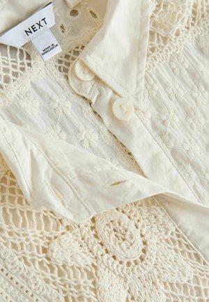 Kremo spalvos marškinių tipo palaidinė su gėlių siuvinėjimu ir nėrinių dizainu. Lygi tekstūra, šalia apykaklės du mygtukai. Medvilnės medžiaga.