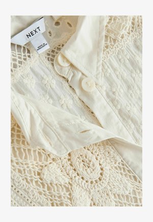 Cremefarbene Hemdbluse mit Blumenstickerei und Spitzen-Design. Glatte Textur mit zwei Knöpfen am Kragen. Baumwollmaterial.