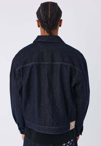 Giacca di denim blu scuro con vestibilità leggermente oversize, chiusura frontale con bottoni e patch con logo sulla parte bassa della schiena. Tessuto strutturato con dettagli di cucitura.