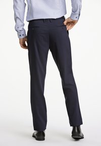 Marineblauwe broek met een getailleerde pasvorm, voorzien van achterzakken met knopen en een gladde stof, gecombineerd met een lichtblauw gestreept shirt en zwarte schoenen.