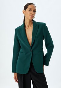 Blazer teal a un bottone con rever a punta, vestibilità su misura e due tasche frontali. Tessuto liscio con una leggera lucentezza, abbinato a pantaloni neri.