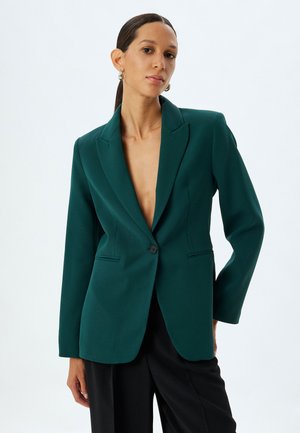 Blazer teal a un bottone con rever a punta, vestibilità su misura e due tasche frontali. Tessuto liscio con una leggera lucentezza, abbinato a pantaloni neri.