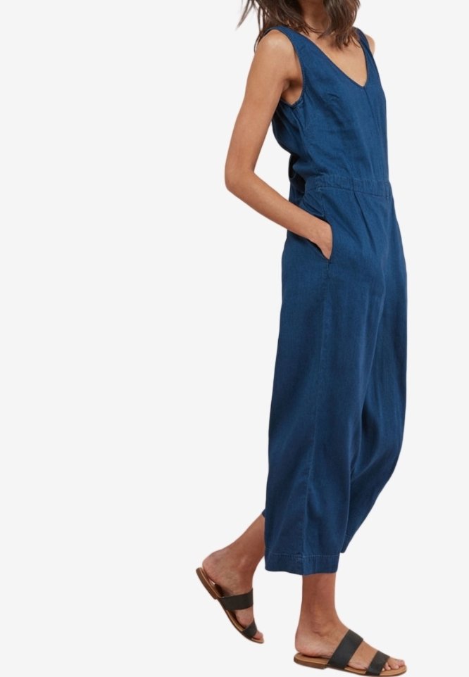 Combinaison en denim bleu avec un col en V, sans manches, poches latérales, jambes larges et coupe décontractée. Portée avec des sandales noires à enfiler.