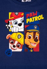 Tre cuccioli sorridenti di Paw Patrol indossano elmetti di salvataggio e uniformi con il testo "PAW PATROL" su uno sfondo blu.