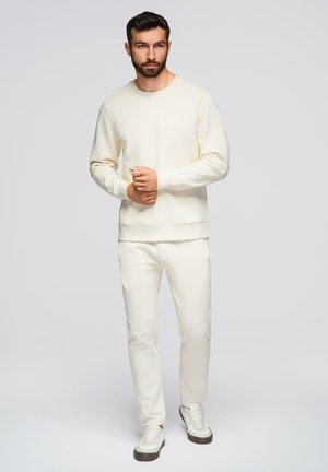 Homme avec barbe portant un sweat-shirt de couleur crème, un pantalon assorti et des baskets blanches, debout devant un fond uni gris clair.