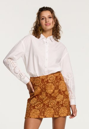 Witte blouse met knoopsluiting en oogjesdetail op de mouwen, gecombineerd met een aansluitende bruine rok met een bloemendessin in gele en oranje tinten.
