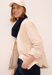 Lys beige kordorjakke med ribbet tekstur, trykknapper og oprullede ærmer. Bæres med et marineblåt tørklæde og mørke jeans.