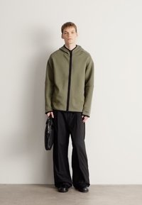 Olívovo zelená mikina na zips s čiernymi akcentmi, doplnená voľnými čiernymi nohavicami a čiernymi topánkami. Model drží čiernu tašku. Jednoduchý dizajn.