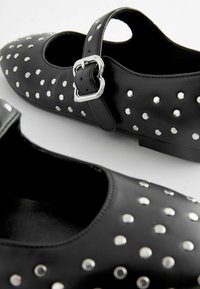 Chaussures en cuir noir avec un bout rond, ornées de clous en argent et d'une sangle avec une boucle en forme de cœur. Texture lisse et semelle plate.