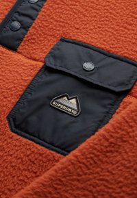 Orange fleece material with a black nylon pocket. The pocket features a snap button and an embroidered mountain logo, adding texture contrast.