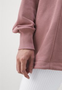 Světle růžová mikina s jemnou texturou; má elastické manžety a mírně oversize střih, spárovaná s bílými elastickými legínami.