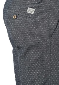 Pantalon gris à motif avec un design subtil en losange, doté d'une poche arrière fermée par un bouton et d'une petite étiquette tissée pour le branding.