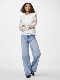 Hvit bluse i blonder med bølgete kanter og volanger på skuldrene, kombinert med brede jeans i blå denim og svarte sko.